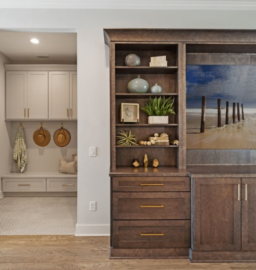 White mudroom cabinetry that includes seating, storage, and hooks in the rear with warm stained wood cabinetry that includes storage, open shelving, and a space for TV/entertainment.